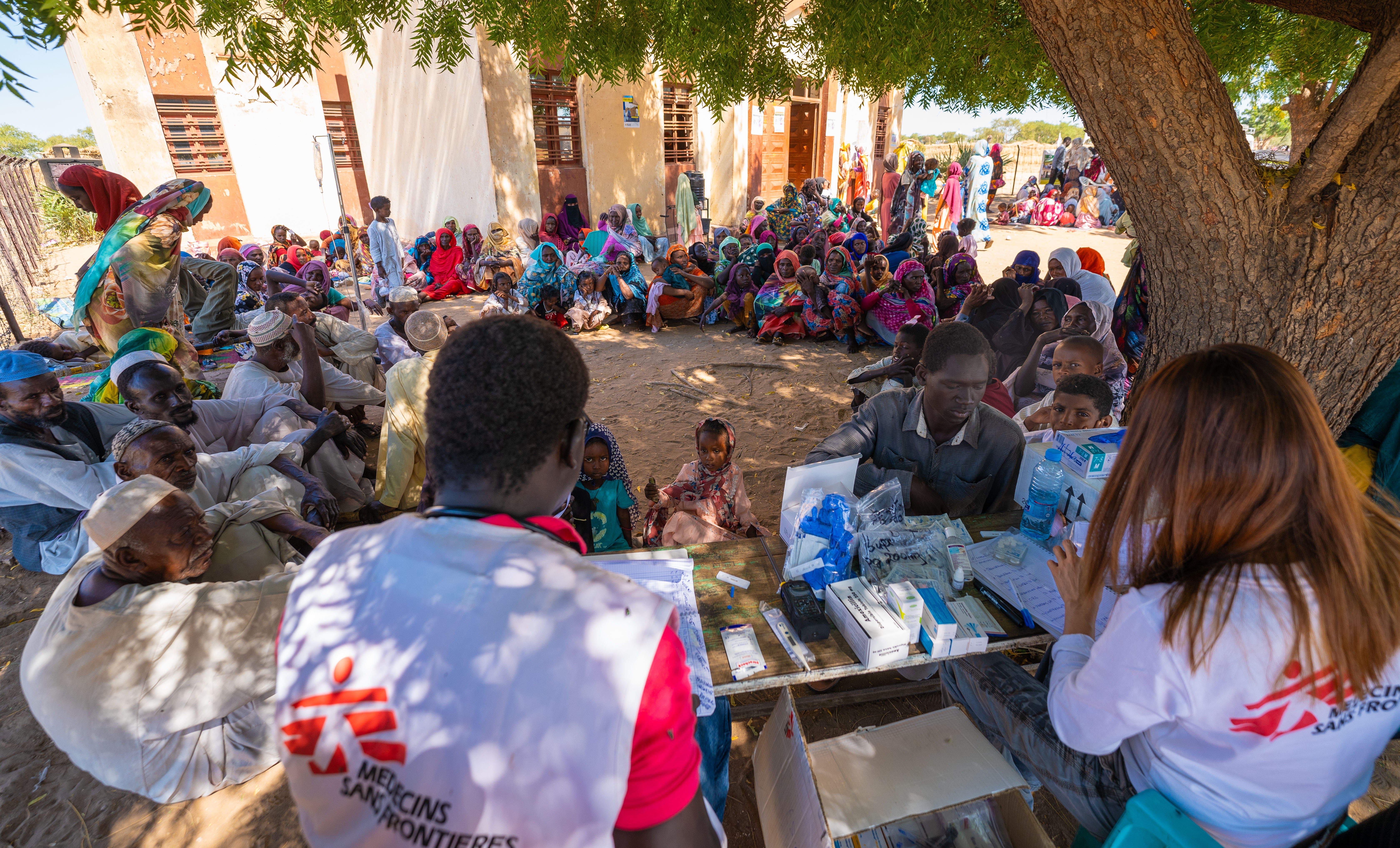 Een menigte repatrianten en vluchtelingen wacht in de rij onder een boom op zorg van het mobiele kliniekteam van AZG in de informele nederzetting Jerbana.© Isaac Buay/MSF