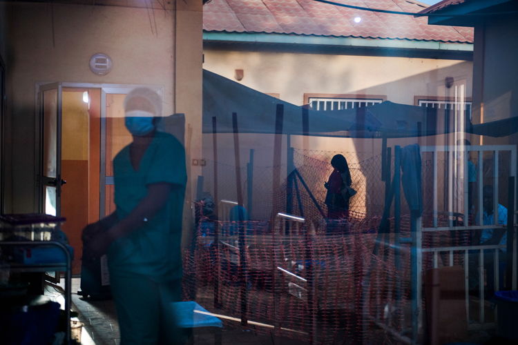 A nurse walks through the room where patients received the diphtheria antitoxin (DAT) while a patient is reflected in the glass door as she talks to visitors at the Centre de Traitement Epidemiologique in Siguiri, Guinea. Visitors are welcome in a limited capacity, having to stand behind a fence to create a two-meter-wide barrier to prevent the spread of diphtheria. MSF has helped treat over 2100 patients as of Jan. 4, 2024, and has been responding to a diphtheria outbreak in the region since mid-August, the likes of which has not been seen in the country for the last 30 years. | Date taken: 12/12/2023 | Photographer: Andrej Ivanov | Location: Guinea