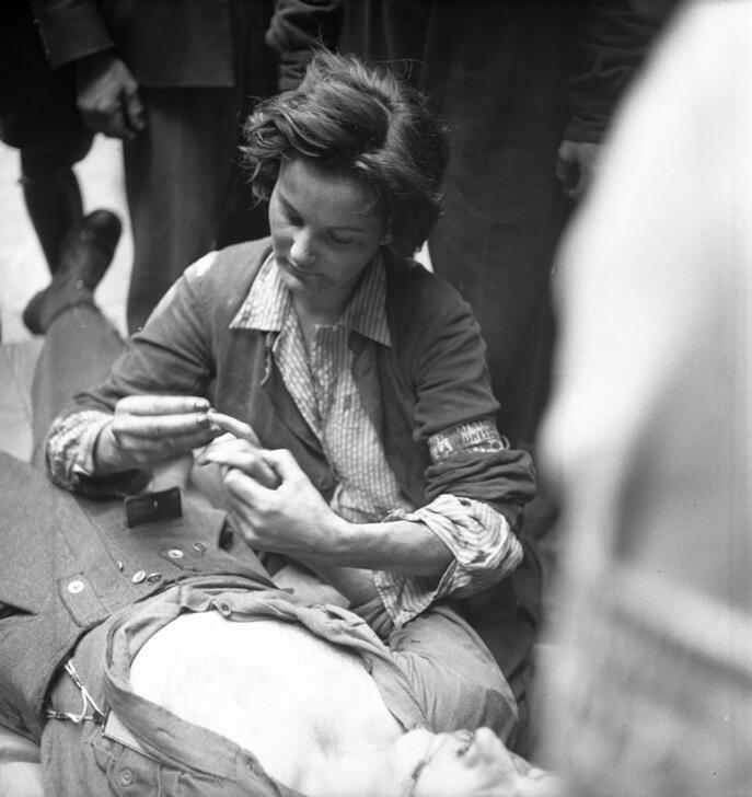 Nurse Anita, holding the hand - removing or replacing the ring - of a wounded man lying on the ground. AKG10620248 © René Zuber / akg-images