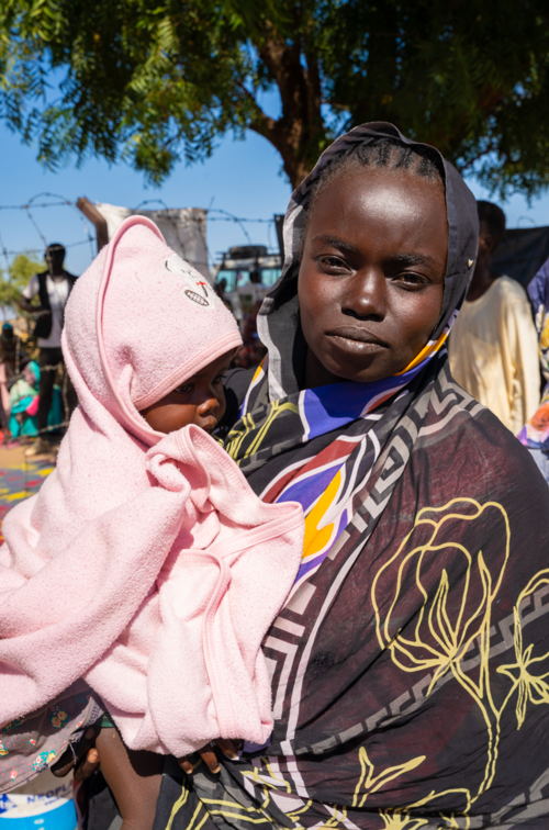 Marriam, 25, houdt haar 1-jarige baby, Iram Taha, vast terwijl ze wacht bij de mobiele kliniek van AZG. Ze vluchtte met een grote groep mensen, terwijl haar familie gestrand blijft in Soedan, zonder contact of updates over hun veiligheid. © Isaac Buay/MSF