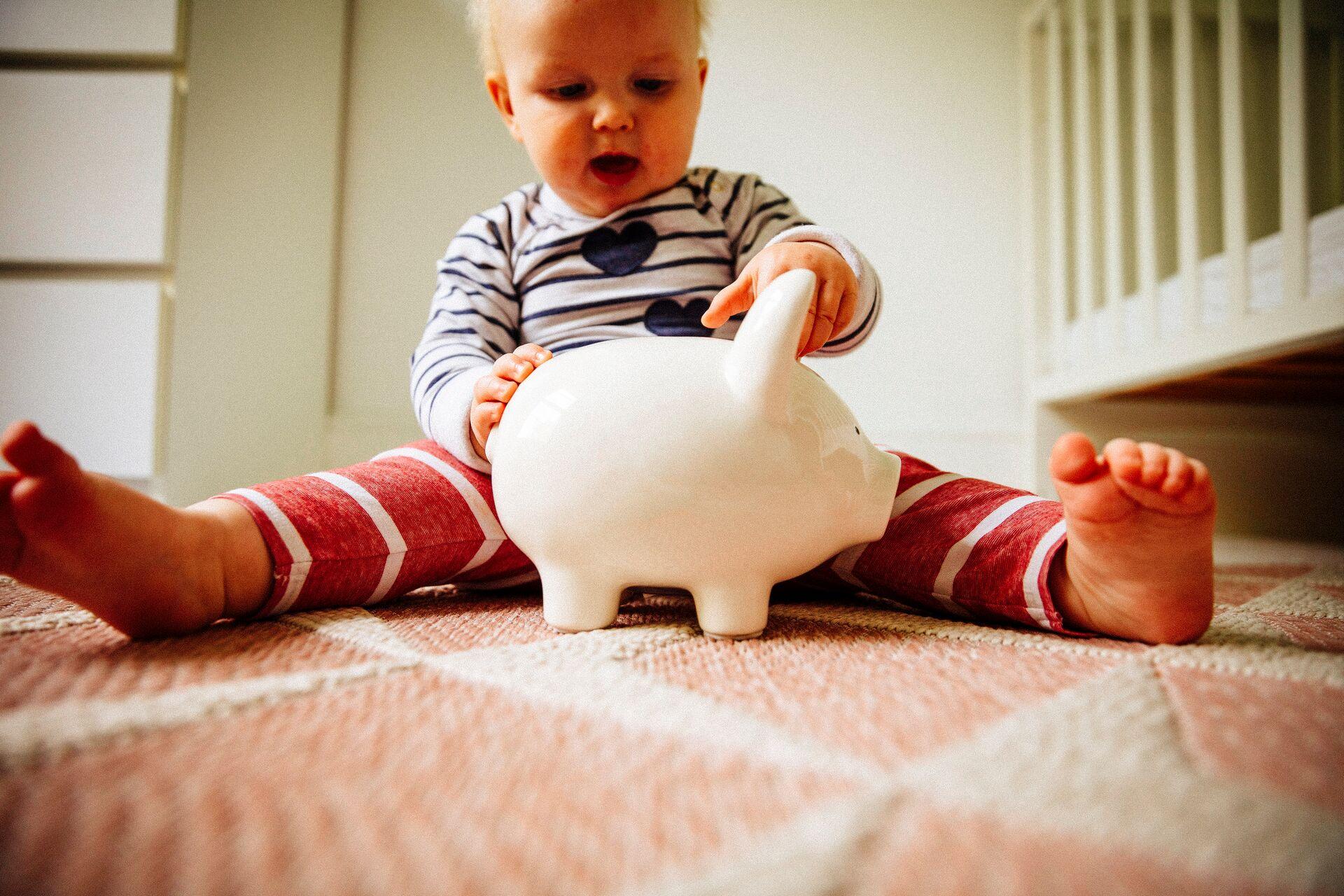 A toddler sits on the floor with a piggy bank, symbolizing changing attitudes toward saving and investing in Belgium