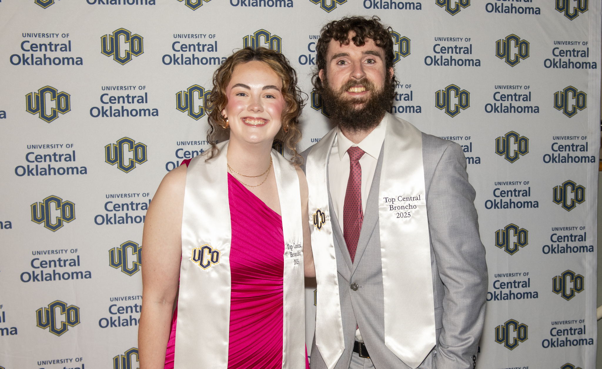 Two UCO students smiling and posing together in front of a UCO backdrop. Two UCO students smiling and posing together in front of a UCO backdrop.