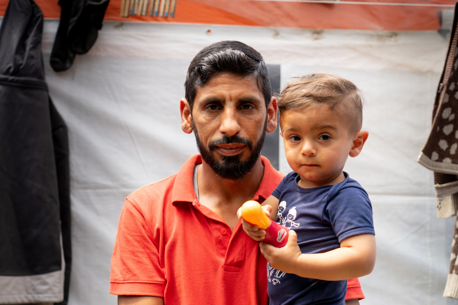 Copyright: Evgenia Chorou/MSF 
Portrait photo of Jaber Alsoudy, his wife and child outside their tent.
Jaber, a 37-year-old man and his wife, 21 years old, left from Syria because of the war, seeking for a safe place and a better future in Europe. During the journey his wife was heavily pregnant. Their son who is now a bit more than 1 year old, was born just before crossing the sea from Turkey.

“We arrived on Samos on November 2019, after multiple failed attempts to cross the sea between Turkey and Greece. We were scared. We spent approximately 9 hours in the sea but finally we made it to Samos and Vathy camp. But there is no safety here. I feel like we are living in a jungle. There is no electricity. There is no good access to water. I feel like we are animals. You constantly have the feeling you are the target. We are a target of the authorities, we are a target of other people inside the camp. We don’t know if something will happen to us by a friend or by an enemy. We don’t feel safe. We are treated like animals.
But despite the circumstances, we are trying to take care of ourselves in these conditions. The long procedure of the asylum application though has a huge impact on us. We are mentally and physically very tired from everything. We are afraid to leave our child outside of the tent because it is not safe and we can’t even go and stay inside the RIC, there it is worse. Here it is safer. There would be no privacy.
It is very hard for us to accept to go to the new camp. We left Syria because we had a lack of dignity and respect for the basic human rights. But what we have been searching in Europe, we have not found it yet. We are not being treated like human beings. We know that the new camp is surrounded by a big fence, that it is in the middle of nowhere, it is an hour away from Vathy. You have to enter and exit that camp at specific times. My wife is attending the mental health clinic of MSF to get psychological care. She is tired and depressed. She never expected that she would need psychiatric medication to cope with life.
We want to have a future. I want to be productive for the society. I want my child to go to school, to have a normal life.” 