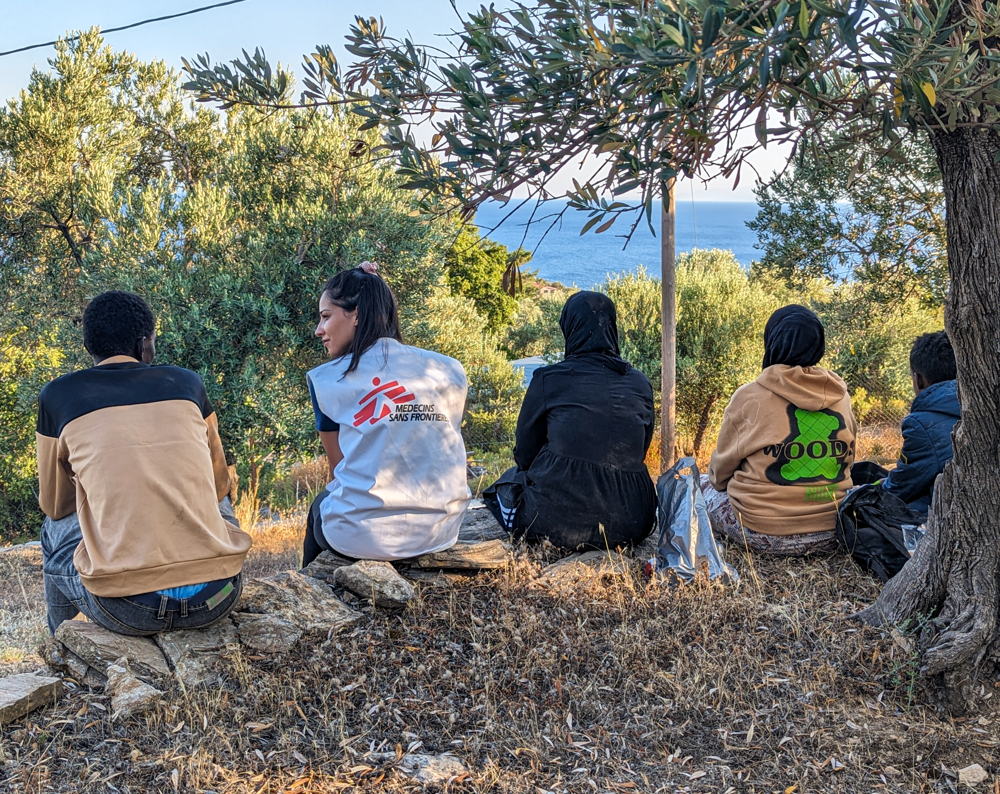 Title: Emergency medical aid activity in Lesvos, Greece Copyright: MSF/Evgenia Chorou Countries: Greece Date taken / Date Recorded: 10 July, 2023 Caption / Description: An MSF team member sits with a group of newly arrived people in Lesvos, who are resting after they have been assisted by MSF teams and provided with food and water. MSF provides emergency assistance – medical and psychological first aid - to people who arrive on the island by boat following notification of people in distress, and thus in need of medical support. Our assistance also includes referrals to hospital by ambulance as well as distribution of food, water and dry clothes. In July 2023, MSF assisted 1,031 people through the emergency medical aid activity, out of whom 26% were children.