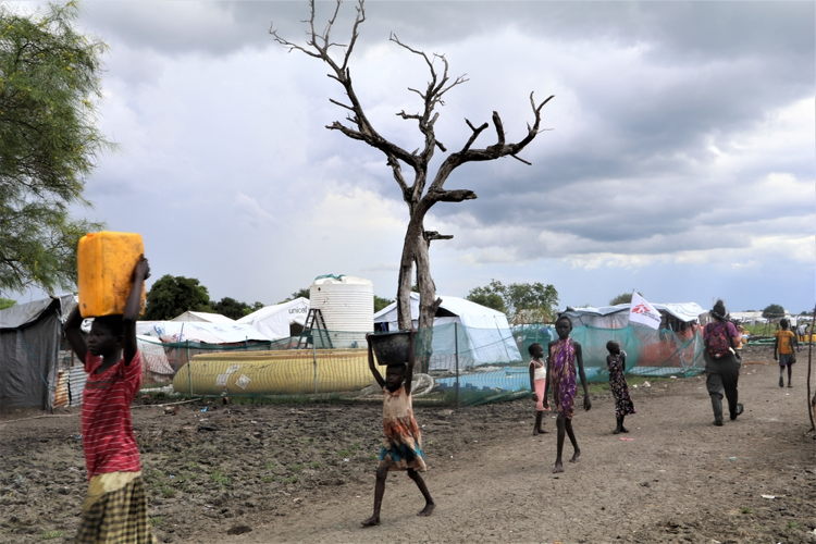 People pass by MSF’s surface water treatment center in Pibor town, the Greater Pibor Administrative Area, South Sudan. MSF has been providing 60 000 liters of drinking water per day to the people of Pibor town after all boreholes became contaminated by floodwaters. This year’s floods are happening against the backdrop of multiple emergencies including COVID-19, increased violence and fighting, a growing economic crisis, and high levels of food insecurity. The needs for medical care are increasing with a sharp rise in malaria cases and fears of outbreaks of other diseases. Photographer: MSF/Tetiana Gaviuk People pass by MSF’s surface water treatment center in Pibor town, the Greater Pibor Administrative Area, South Sudan. MSF has been providing 60 000 liters of drinking water per day to the people of Pibor town after all boreholes became contaminated by floodwaters. This year’s floods are happening against the backdrop of multiple emergencies including COVID-19, increased violence and fighting, a growing economic crisis, and high levels of food insecurity. The needs for medical care are increasing with a sharp rise in malaria cases and fears of outbreaks of other diseases. Photographer: MSF/Tetiana Gaviuk