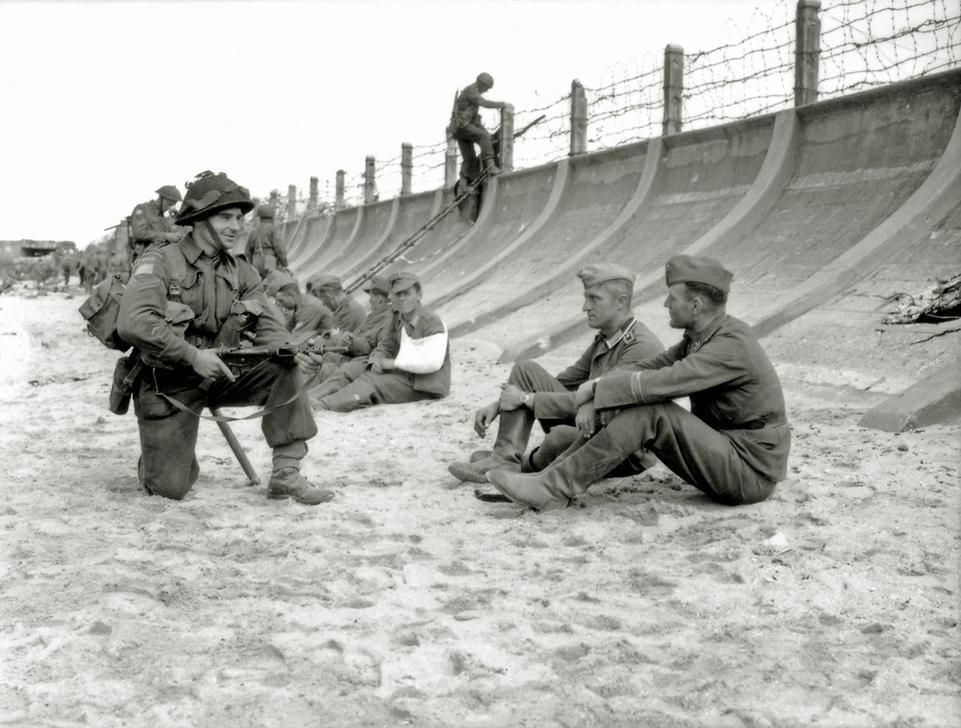 Corporal Victor Deblois from the 3rd Canadian Infantry Division guarding two German prisoners at the foot of the concrete dam at Juno Beach. In the background other prisoners are sitting along the concrete dam in the near Widerstandsnest. AKG2492488 © akg-images
