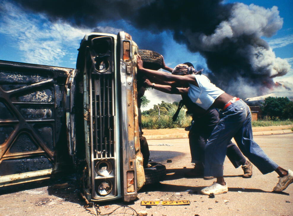People push over a burned-out car, as smoke billows from a building in the background, South Africa. AKG10784707 © akg-images / Ken Oosterbroek / Independent Photographers / african.pictures