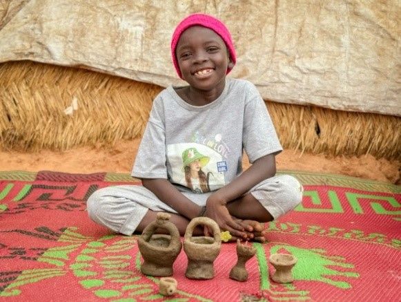 Rayan, 7, is one of the tens of thousands of children whose families have had to flee the extreme violence of the war in Sudan. She proudly poses in front of little cups and tea pots she makes from mud she picks up after the rain. “When I was still going to school in Sudan”, she explains, “my favourite moment was the end of the class, when the teacher was giving us a toy tea set to play with.” In the transit camp, Rayan no longer goes to school, “but,” she says “I make tea sets of my own, so I can keep playing with the other kids”.