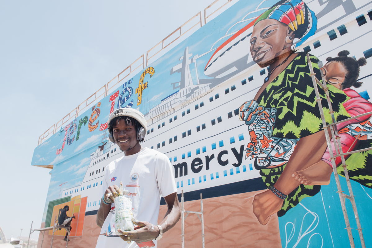 Ahmadou Moustapha Tounkara (connu sous le nom de Lartis Ka), artiste sénégalais, devant la fresque qu'il peint pour l'arrivée du Global Mercy sur le quai supérieur du port de Dakar.