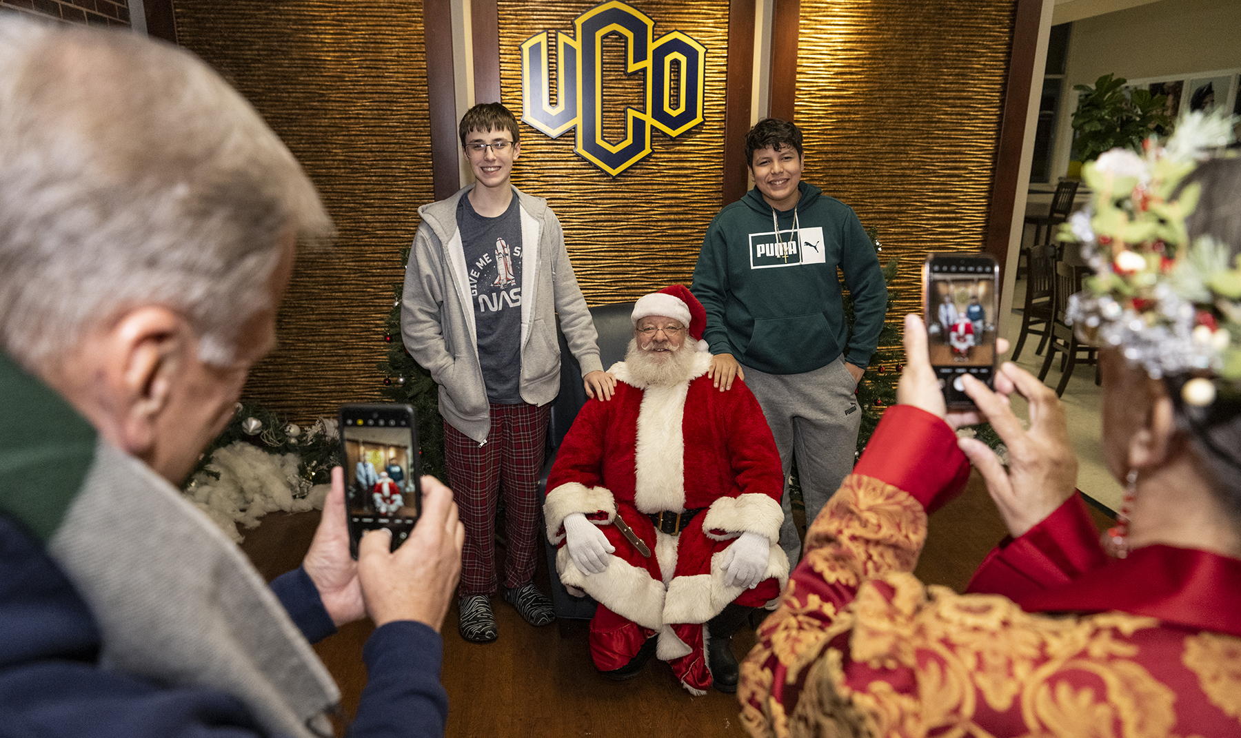 Two people posing with Santa for a photo as two people use phones to take the photo. Two people posing with Santa for a photo as two people use phones to take the photo. 