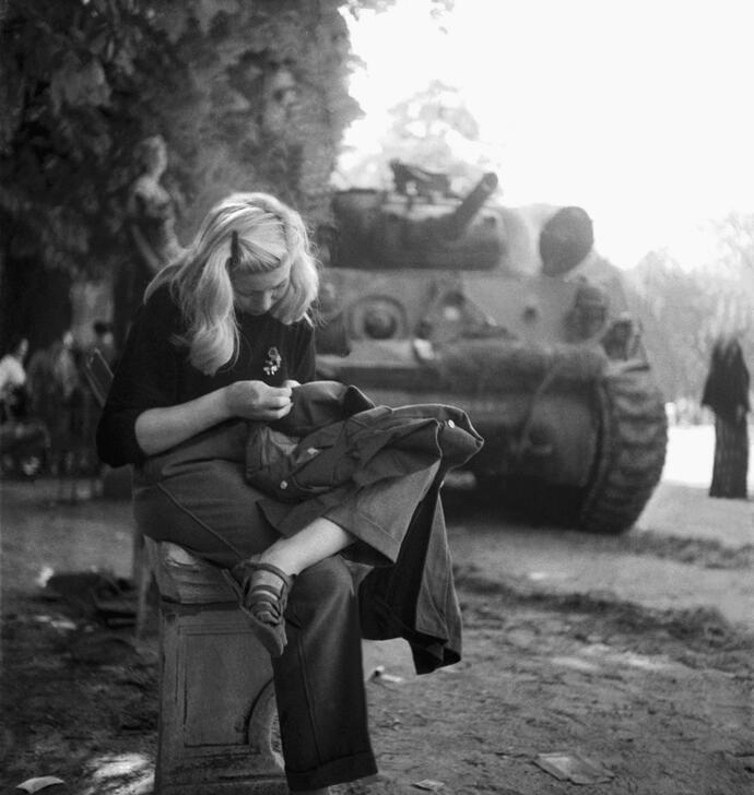 A young woman sitting on a bench on a boulevard, close to a tank, appears to be sewing a military uniform jacket. AKG10778562 © René Zuber / akg-images
